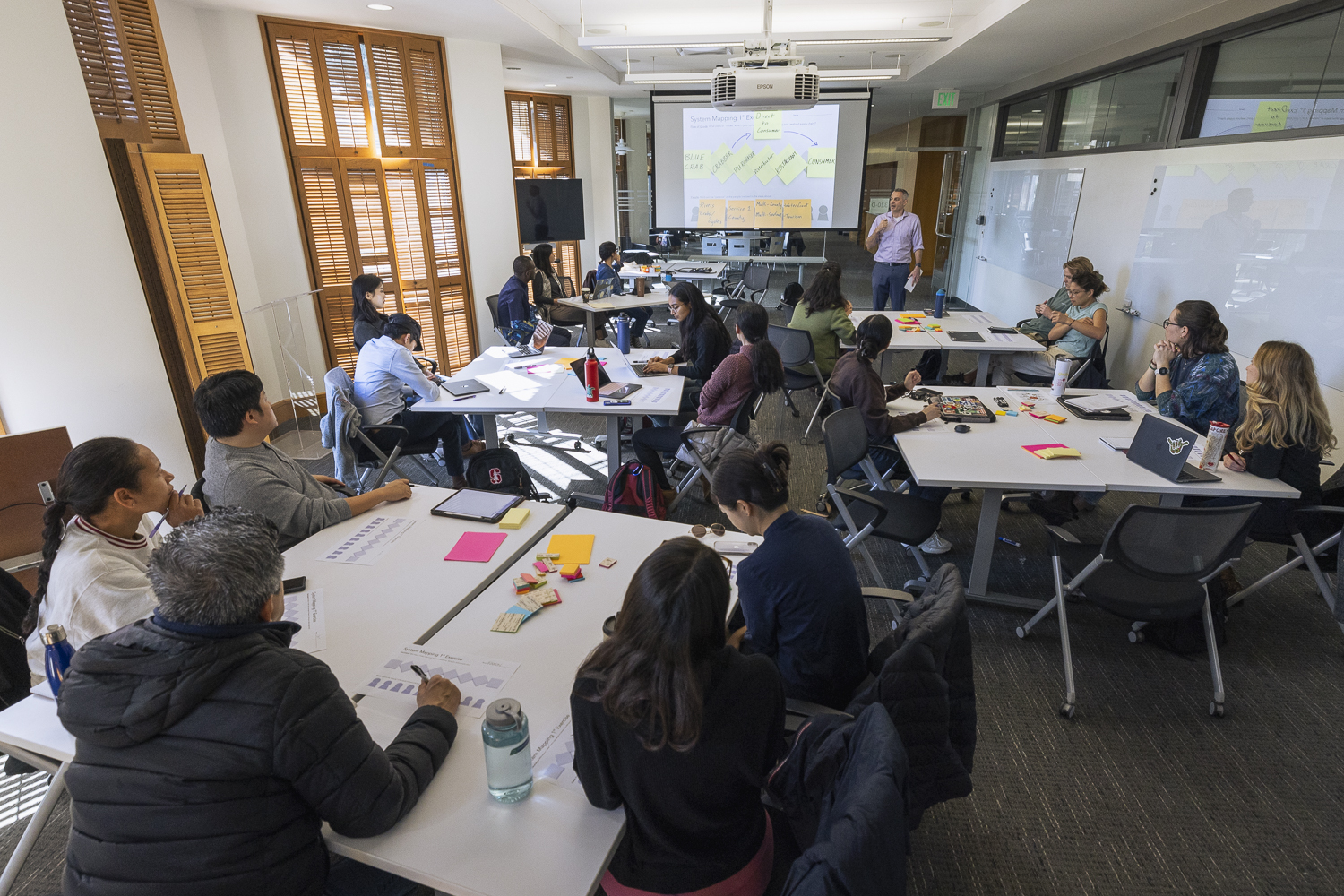 A large classroom full of worktables with students sit, busy taking notes and listening to a lecturer who stands at the front of the room, next to a large screen. Image credit: Andrew Brodhead