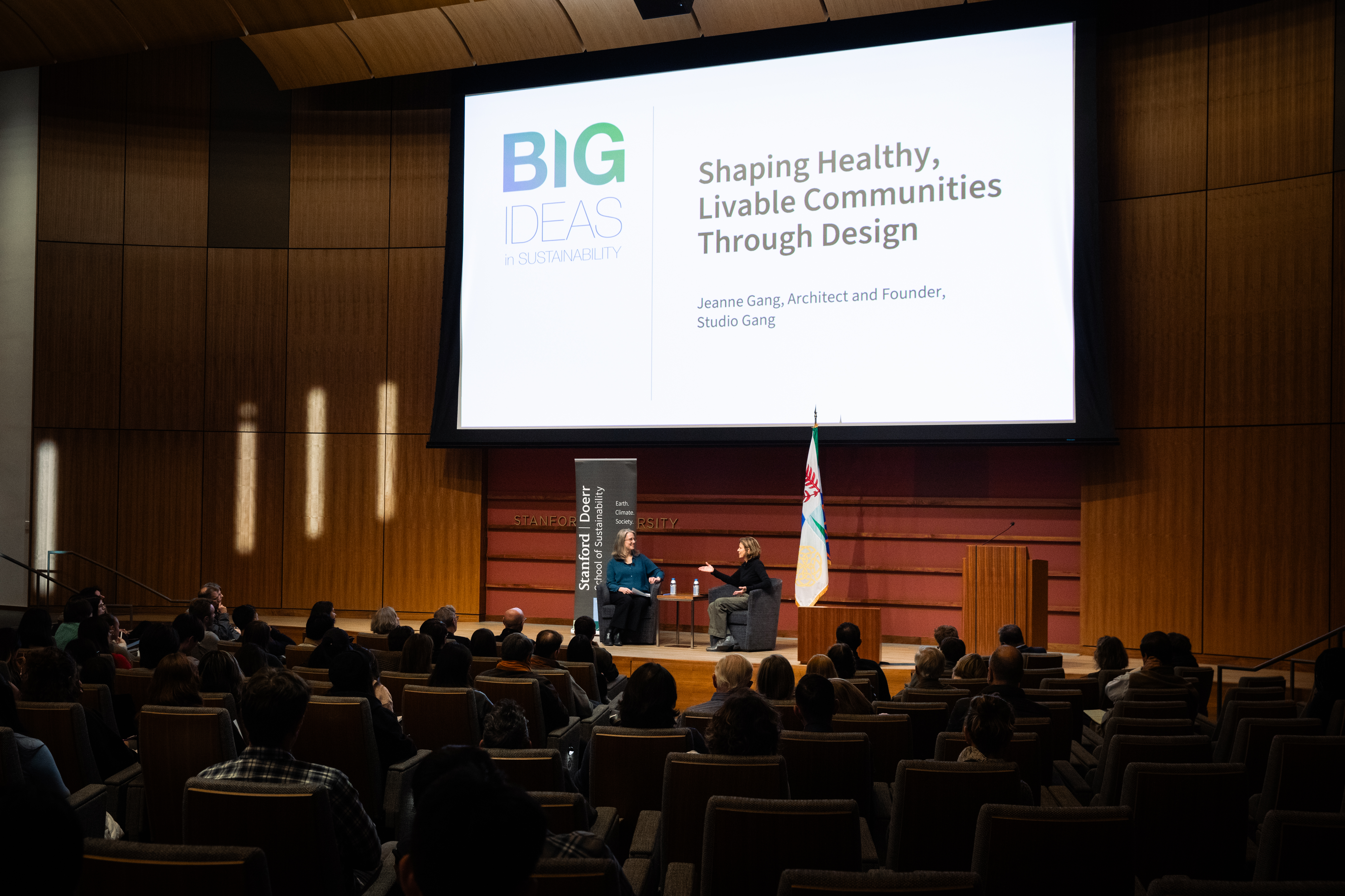 Jeanne Gang and Sarah Billington sit on a low stage under a large screen displaying the Big Ideas event title above them and large audience in foreground. Image credit: Patrick Beaudouin