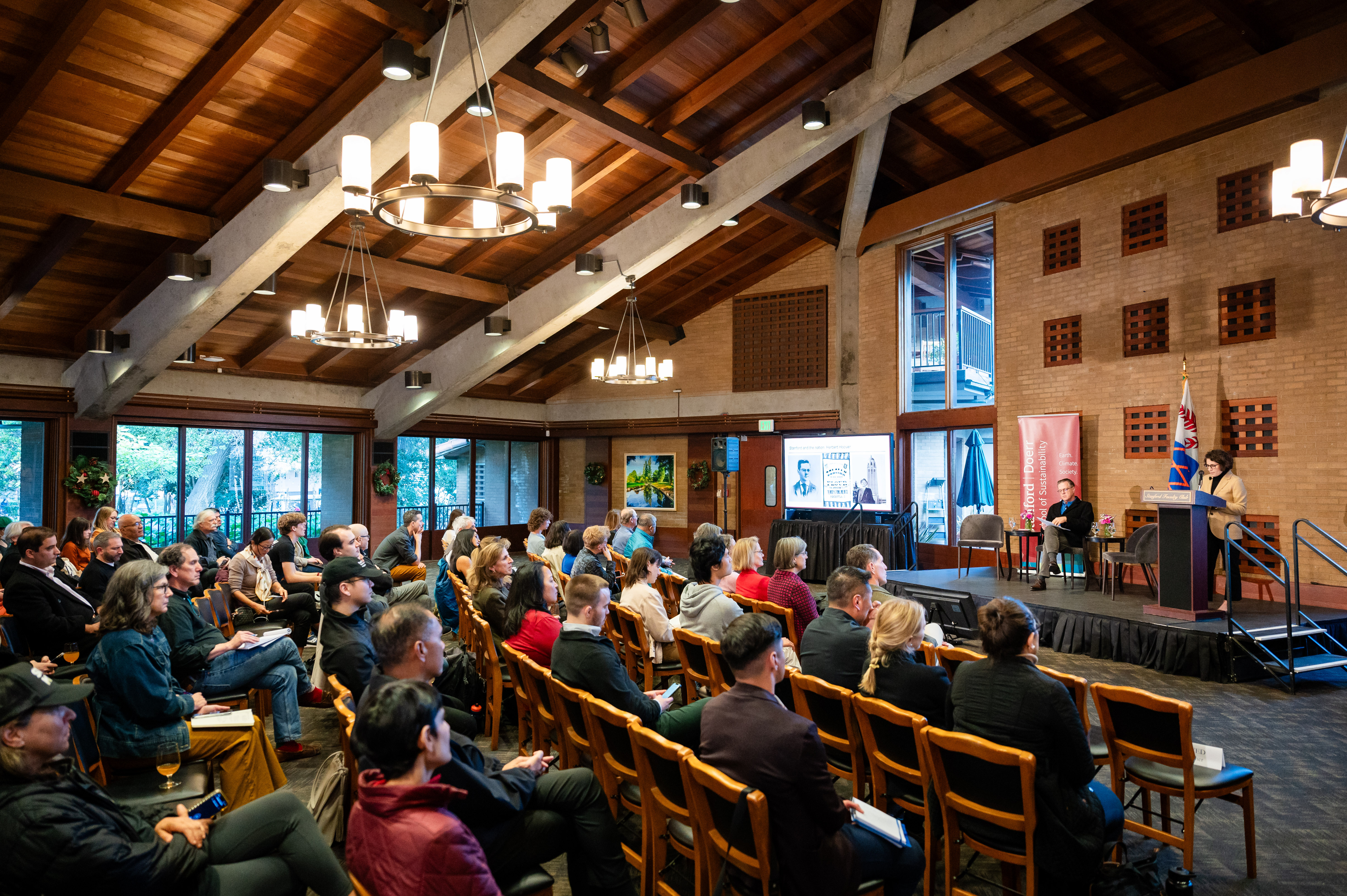 A large audience sits in a spacious room listening to a panel discussion on a low stage, with a large screen to the left of stage. Image credit: SF Photo Agency