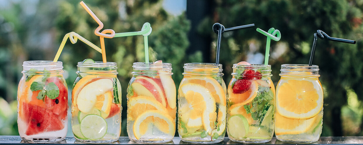 Six mason jars filled with juice, fresh watermelon, lime, apple, orange, strawberries, mint, and topped with colorful bendy straws set outdoors on a sunny day