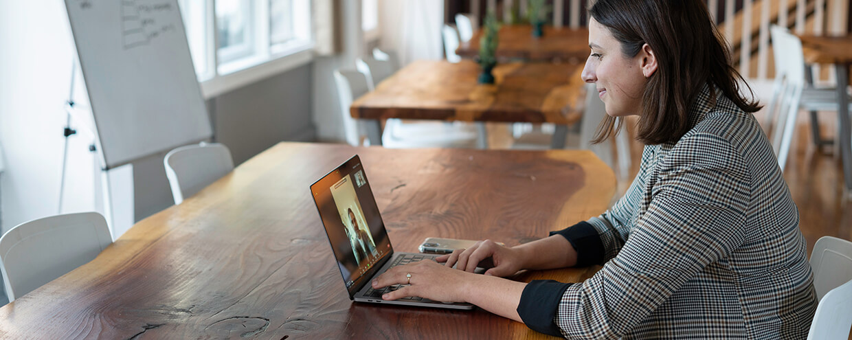 A woman in business attire sits at a hardwood table in front of her laptop. She smiles at the other people in the online meeting she’s attending.